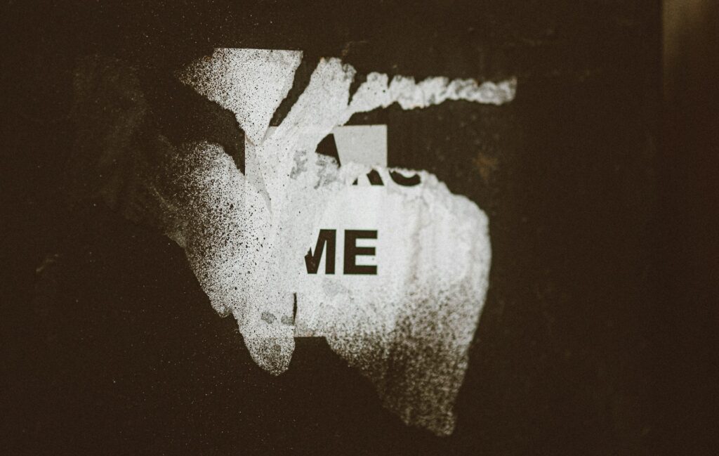 Distressed white map silhouette with the letters ‘ME’ on a dark, textured background, symbolizing fractured identity.