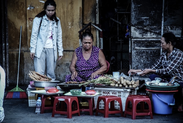 Người phụ nữ ngồi bán đồ ăn trên vỉa hè với nhiều chiếc ghế nhựa đỏ thấp bao quanh – hình ảnh đặc trưng của văn hóa ẩm thực đường phố Việt Nam.